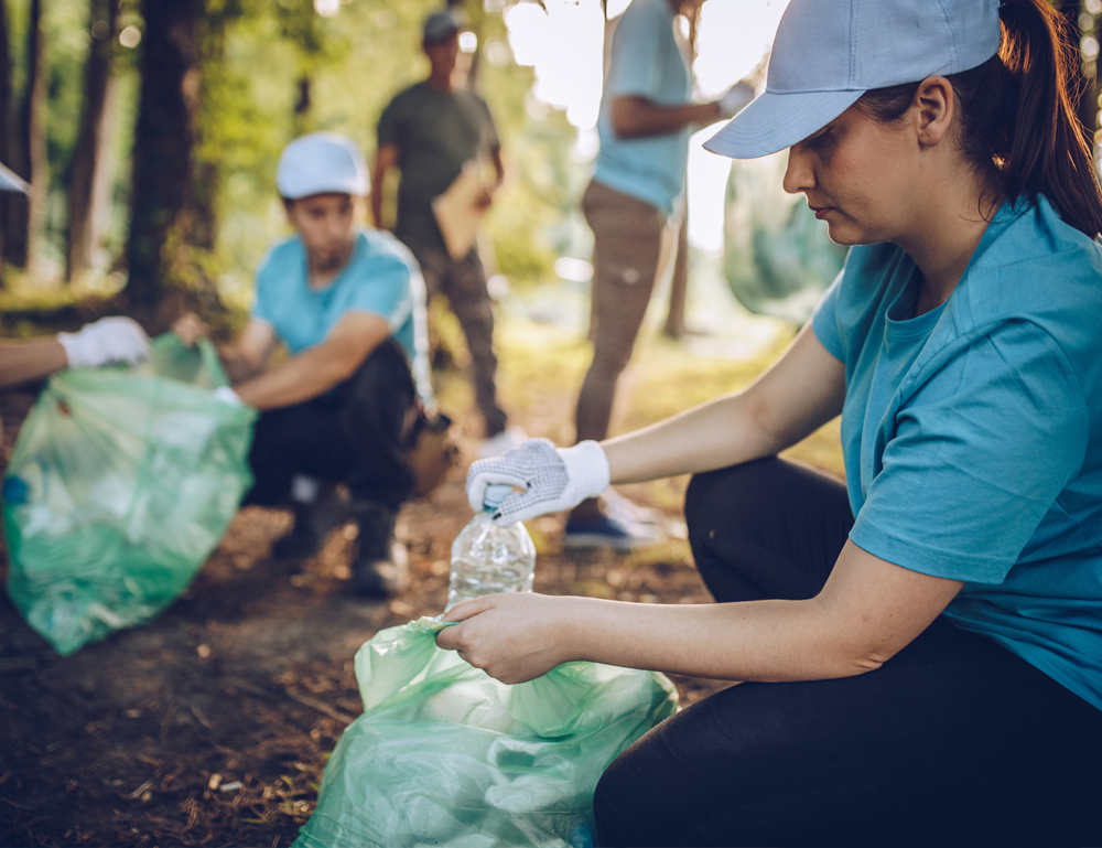 People wearing gloves pick up empty plastic bottles from the ground and put them into large green plastic bags.