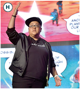 Gabby Rivera raises her right arm and looks to the front. Some big-sized cartoons are showing behind her.